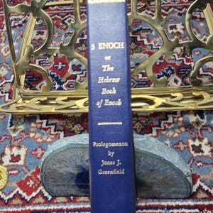 May include: A blue hardcover book titled "3 Enoch or The Hebrew Book of Enoch" by Jonas J. Greenfield. The book is propped up by a blue and gray stone bookend. The book has gold lettering and is in front of a gold metal book stand.