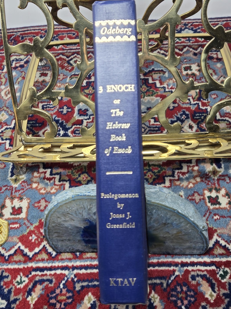 May include: A blue hardcover book titled "3 Enoch or The Hebrew Book of Enoch" by Jonas J. Greenfield. The book is propped up by a blue and gray stone bookend. The book has gold lettering and is in front of a gold metal book stand.