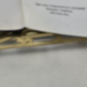 Puede incluir: Imagen borrosa de un libro con texto y un elemento decorativo dorado. El texto es parcialmente visible, con l&iacute;neas de texto y algunas palabras. El elemento decorativo parece ser un dise&ntilde;o de filigrana.