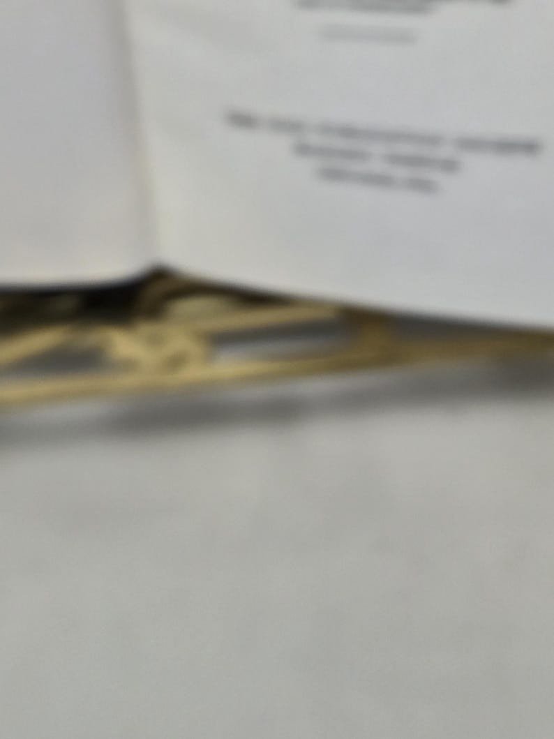 Puede incluir: Imagen borrosa de un libro con texto y un elemento decorativo dorado. El texto es parcialmente visible, con l&iacute;neas de texto y algunas palabras. El elemento decorativo parece ser un dise&ntilde;o de filigrana.
