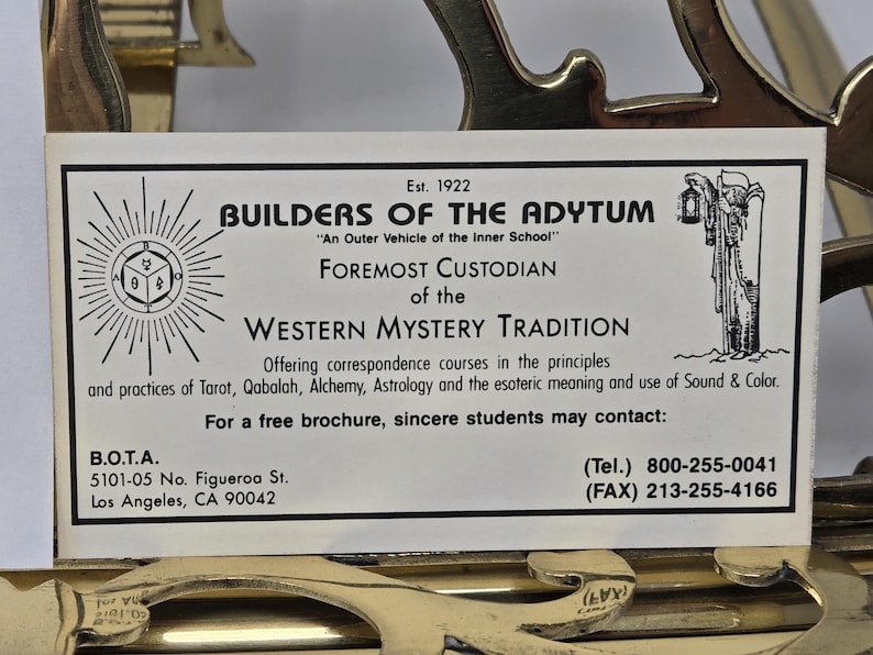 Puede incluir: Una tarjeta de presentaci&oacute;n de Builders of the Adytum, establecida en 1922. La tarjeta presenta informaci&oacute;n sobre la Tradici&oacute;n del Misterio Occidental, incluyendo Tarot, C&aacute;bala y Alquimia. La tarjeta est&aacute; sobre un elemento decorativo dorado.