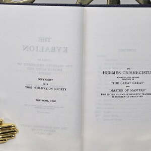 Puede incluir: Libro abierto con texto, incluyendo "THE KYBALION" y "COPYRIGHT 1912 YOGI PUBLICATION SOCIETY." Dedicado a Hermes Trismegisto, "THE GREAT GREAT" y "MASTER OF MASTERS." Se ve un sujetalibros dorado en forma de mano.