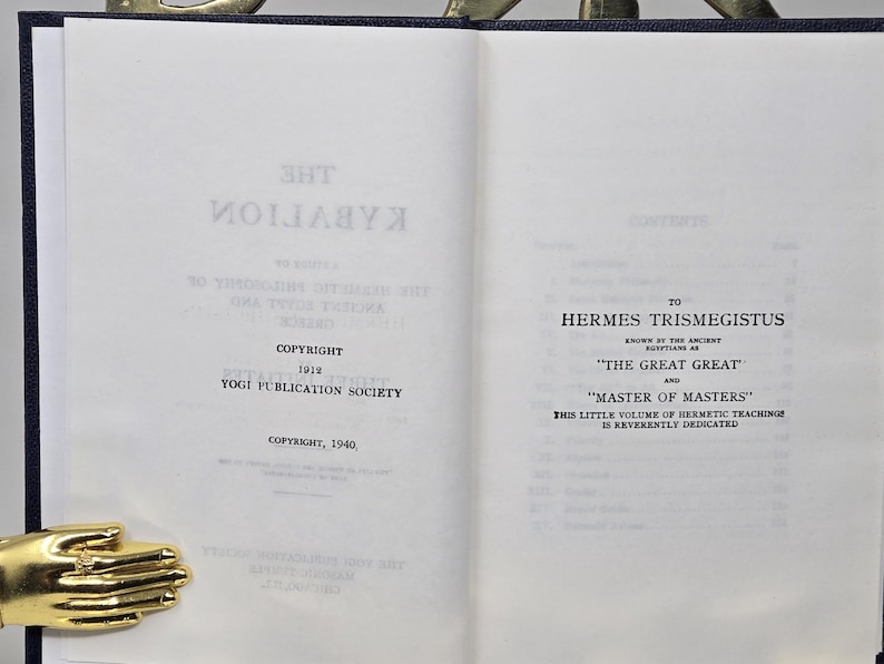Puede incluir: Libro abierto con texto, incluyendo "THE KYBALION" y "COPYRIGHT 1912 YOGI PUBLICATION SOCIETY." Dedicado a Hermes Trismegisto, "THE GREAT GREAT" y "MASTER OF MASTERS." Se ve un sujetalibros dorado en forma de mano.