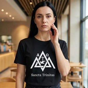 May include: Black t-shirt with a white geometric design featuring a dove, cross, and Greek letters. The text "Sancta Trinitas" is below the design. The person is wearing black jeans.