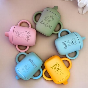 Pode incluir: Cinco copos de aprendizagem de silicone coloridos com nomes personalizados, incluindo "Adalind", "Angel", "Luke", "Taniqua" e "Bella". Cada copo apresenta um design de animal diferente, como um caracol, um coelho, um urso, um le&atilde;o e uma abelha.