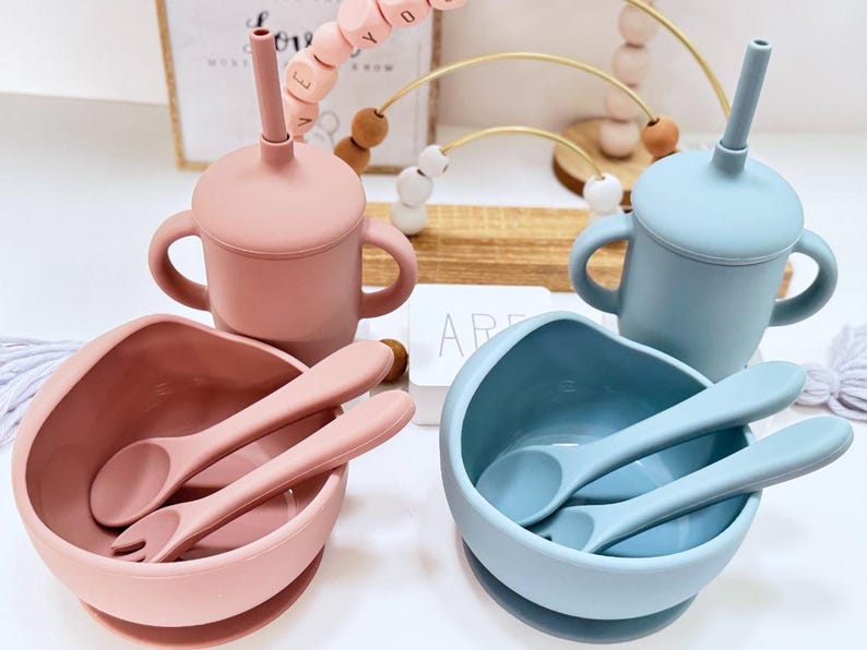 May include: A baby feeding set in pink and blue. The set includes two sippy cups with handles and straws, two bowls with spoons and forks, and a wooden bead toy. The items are made of silicone.