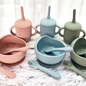 May include: A collection of baby feeding items in pastel shades. The set features three sippy cups with handles and straws, three bowls with spoons, and three forks. The items are in pink, blue, and green.