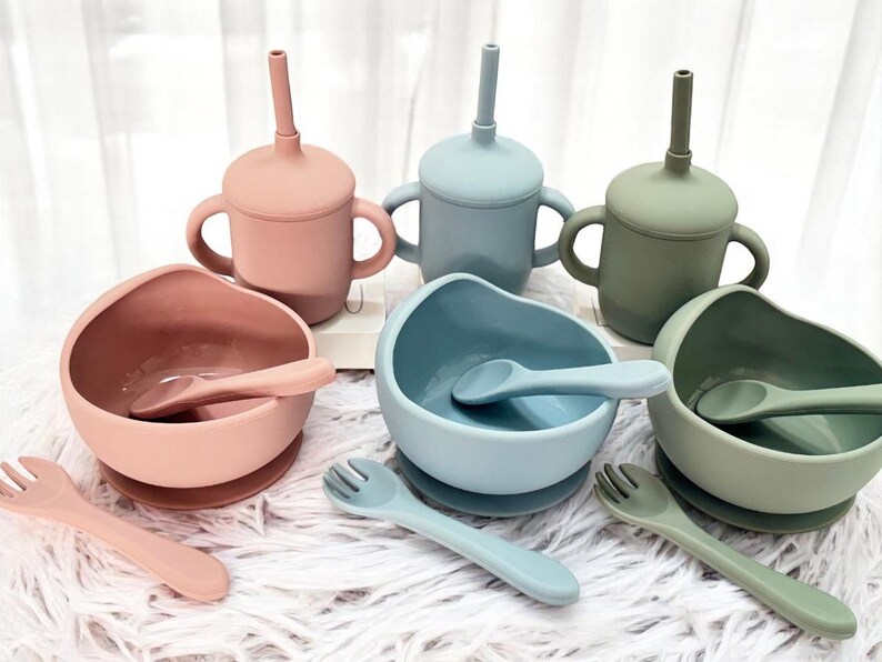 May include: A collection of baby feeding items in pastel shades. The set features three sippy cups with handles and straws, three bowls with spoons, and three forks. The items are in pink, blue, and green.