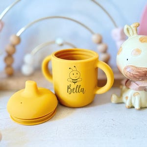 Pode incluir: Copo de treinamento de silicone amarelo com duas al&ccedil;as e uma tampa. O copo tem um desenho de abelha e o nome "Bella" impresso.