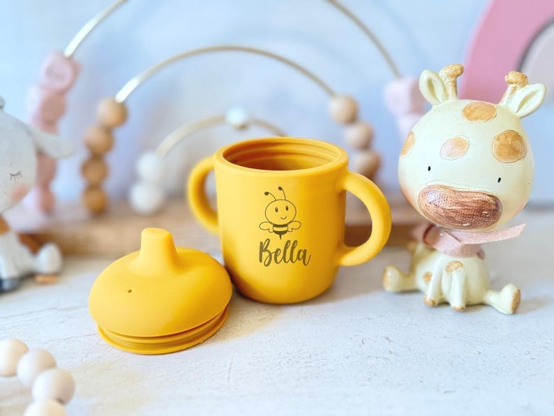 Pode incluir: Copo de treinamento de silicone amarelo com duas al&ccedil;as e uma tampa. O copo tem um desenho de abelha e o nome "Bella" impresso.