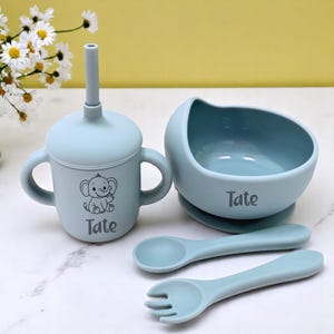 May include: A light blue baby feeding set comprising a cup with handles, a bowl, and two utensils. The cup and bowl are personalized with the name "Tate" and an elephant graphic. The set is made of silicone.