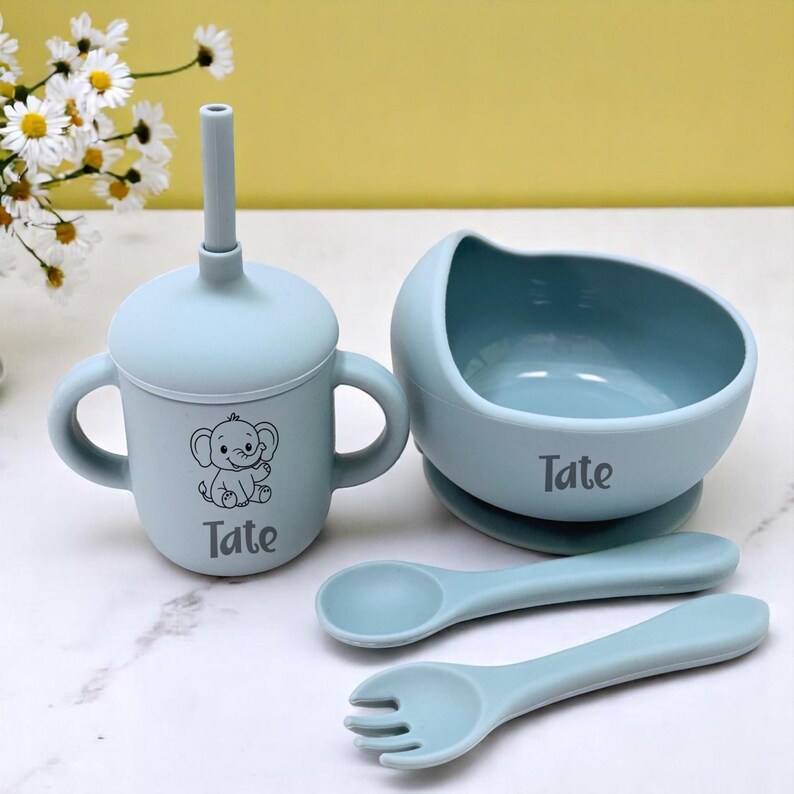 May include: A light blue baby feeding set comprising a cup with handles, a bowl, and two utensils. The cup and bowl are personalized with the name "Tate" and an elephant graphic. The set is made of silicone.