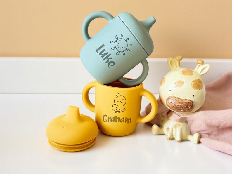 Pode incluir: Duas canecas de silicone com al&ccedil;as, uma azul e uma amarela. A caneca azul tem um desenho de sol e o texto "Luke" nela. A caneca amarela tem um desenho de pintinho e o texto "Graham" nela. Tamb&eacute;m &eacute; mostrado uma tampa de silicone amarela.