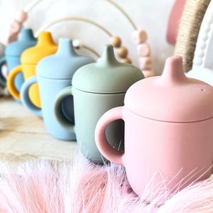 Pode incluir: Cinco copos de silicone com tampas em diferentes cores: azul, amarelo, azul claro, verde e rosa. Os copos t&ecirc;m duas al&ccedil;as e est&atilde;o alinhados numa superf&iacute;cie de madeira.