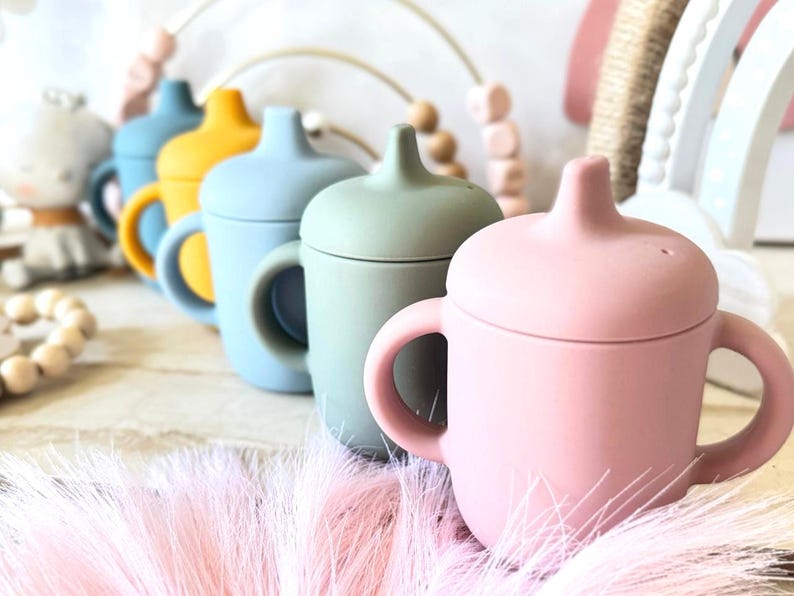 Pode incluir: Cinco copos de silicone com tampas em diferentes cores: azul, amarelo, azul claro, verde e rosa. Os copos t&ecirc;m duas al&ccedil;as e est&atilde;o alinhados numa superf&iacute;cie de madeira.