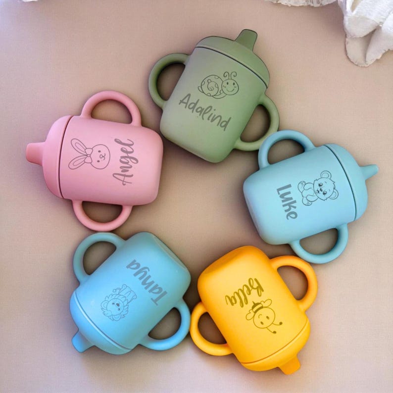 Pode incluir: Cinco copos de aprendizagem de silicone coloridos com nomes personalizados, incluindo "Adalind", "Angel", "Luke", "Taniqua" e "Bella". Cada copo apresenta um design de animal diferente, como um caracol, um coelho, um urso, um le&atilde;o e uma abelha.