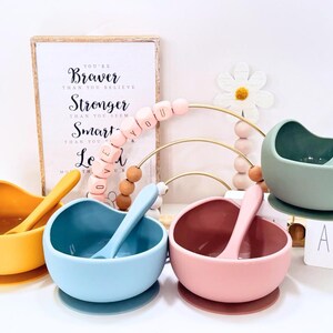 May include: Four silicone baby bowls in yellow, blue, pink, and green, each with a matching spoon. A framed print with the words "You're Braver Than You Believe" is in the background, along with a decorative bead arch.