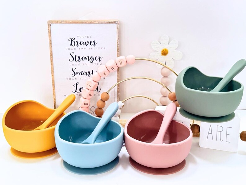 May include: Four silicone baby bowls in yellow, blue, pink, and green, each with a matching spoon. A framed print with the words "You're Braver Than You Believe" is in the background, along with a decorative bead arch.