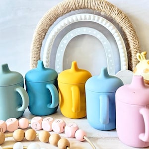 Pode incluir: Cinco copos de beb&eacute; de silicone em diferentes cores: azul-esverdeado, azul, amarelo, azul claro e rosa. Os copos t&ecirc;m uma tampa e uma pega. Est&atilde;o dispostos em fila sobre uma superf&iacute;cie branca.