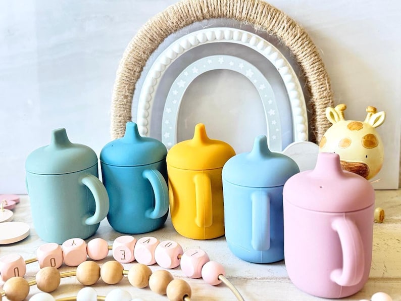 Pode incluir: Cinco copos de beb&eacute; de silicone em diferentes cores: azul-esverdeado, azul, amarelo, azul claro e rosa. Os copos t&ecirc;m uma tampa e uma pega. Est&atilde;o dispostos em fila sobre uma superf&iacute;cie branca.