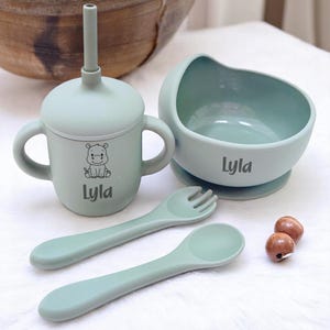 May include: A light green baby feeding set including a cup, bowl, spoon, and fork. The cup has handles and a straw, and features a hippo graphic and the name "Lyla". The bowl and cup have the name "Lyla" printed on them.