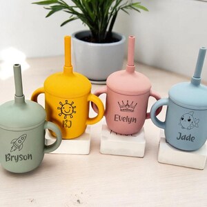 May include: Four pastel-coloured silicone sippy cups with matching straws and handles. The cups are green, yellow, pink, and blue, each personalised with a name and illustration: a rocket, a sun, a crown, and a fish.