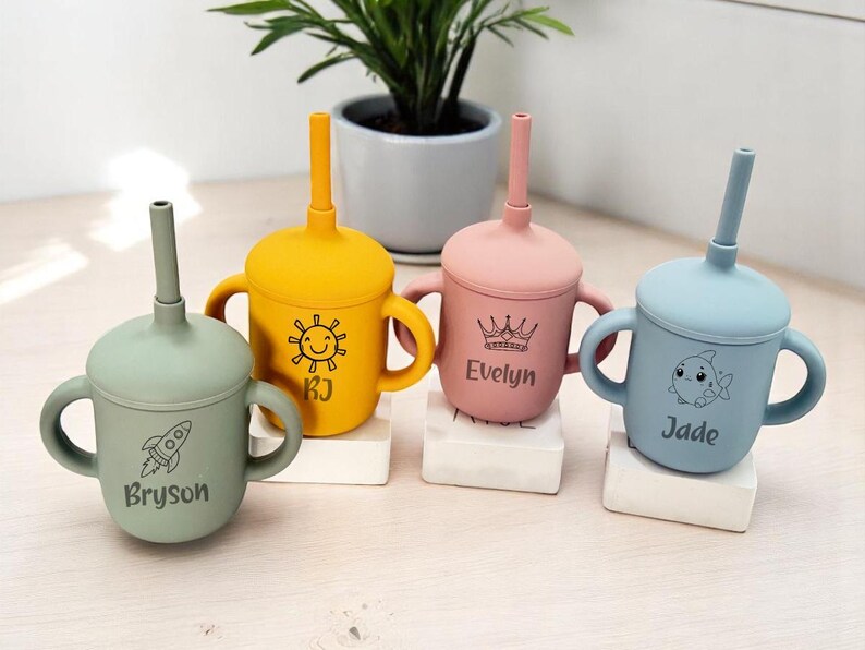 May include: Four pastel-coloured silicone sippy cups with matching straws and handles. The cups are green, yellow, pink, and blue, each personalised with a name and illustration: a rocket, a sun, a crown, and a fish.