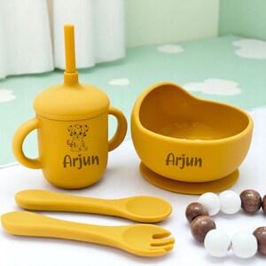 May include: A mustard yellow baby feeding set comprising a sippy cup with a straw, a bowl, a spoon, and a fork. The cup and bowl have the name "Arjun" printed on them. A wooden bead bracelet is also included.