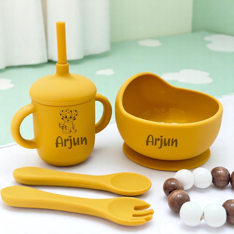 May include: A mustard yellow baby feeding set comprising a sippy cup with a straw, a bowl, a spoon, and a fork. The cup and bowl have the name "Arjun" printed on them. A wooden bead bracelet is also included.