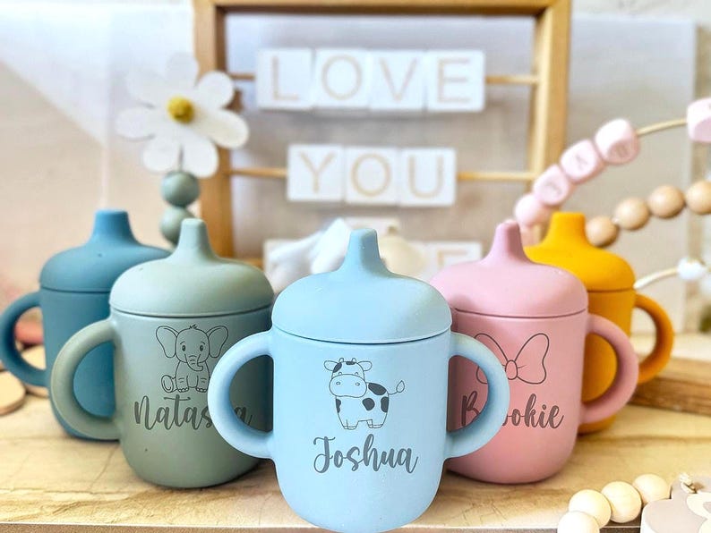 Pode incluir: Quatro copos de treinamento de silicone personalizados com tampas. Os copos s&atilde;o azuis, verdes, rosa e amarelos. Cada copo tem um design de animal diferente e um nome na frente. Os nomes s&atilde;o Joshua, Natas, Brookie e um quarto nome que n&atilde;o &eacute; vis&iacute;vel.