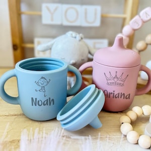 Pode incluir: Duas canecas de silicone personalizadas com al&ccedil;as. A caneca azul tem um desenho de um menino e o nome "Noah" impresso nela. A caneca rosa tem uma coroa e o nome "Ariana" impresso nela.
