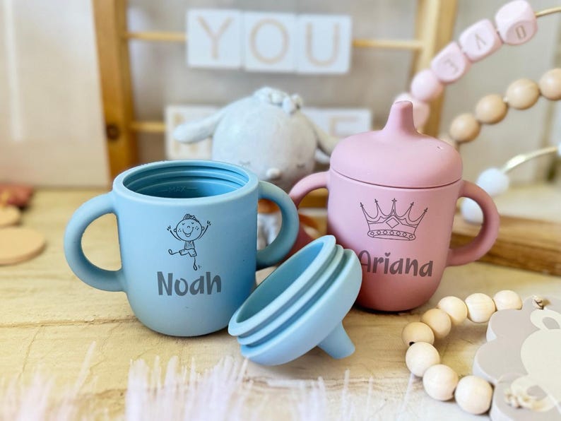 Pode incluir: Duas canecas de silicone personalizadas com al&ccedil;as. A caneca azul tem um desenho de um menino e o nome "Noah" impresso nela. A caneca rosa tem uma coroa e o nome "Ariana" impresso nela.