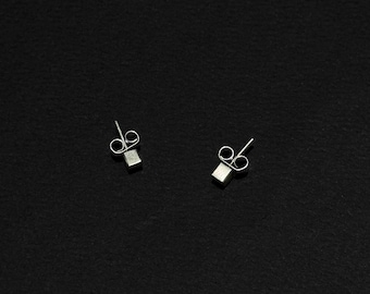 Puede incluir: Un par de pendientes de botón cuadrados y pequeños de plata. Los pendientes se presentan sobre un fondo negro, resaltando su diseño minimalista. También son visibles los postes y los cierres de mariposa.