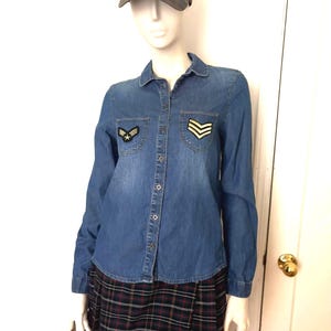May include: A denim shirt with patches and a plaid skirt are displayed on a mannequin. The shirt has a button-down front and long sleeves. A camouflage baseball cap sits atop the mannequin's head.