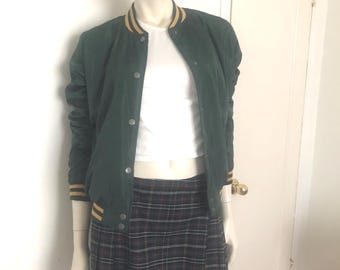 May include: A mannequin wearing a dark green bomber jacket with yellow stripes on the collar and cuffs, a white top, and a plaid skirt. A camouflage baseball cap completes the outfit.