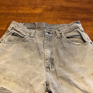 Könnte beinhalten: Verblasste graue Jeans mit einem Used-Look. Die Jeans haben einen Knopfverschluss, Vordertaschen und ein gerissenes Knie. Es gibt einige Flecken auf der Vorderseite. Ein klassischer Jeans-Stil.