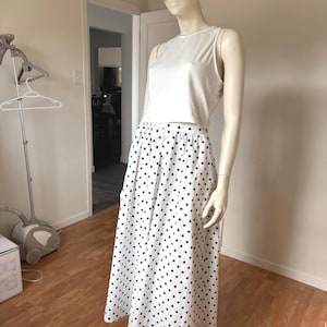Puede incluir: Una camiseta sin mangas blanca y una falda midi blanca con lunares azul marino se exhiben en un maniquí. Una gorra de béisbol de camuflaje se usa en la cabeza del maniquí. El atuendo está diseñado en un maniquí.