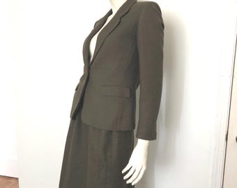 May include: Olive green skirt suit with a matching blazer and a straw hat with a black band. The suit features a classic tailored design. The hat is a fedora style. The outfit is displayed on a mannequin.