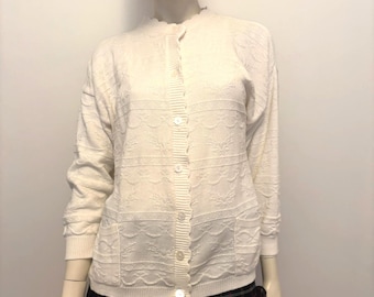 Puede incluir: Rebeca color crema con cuello festoneado y cierre de botones. El suéter de manga larga presenta un patrón floral texturizado y dos bolsillos delanteros. Un sombrero de paja con una cinta negra está encima.