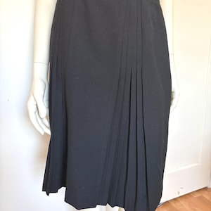 May include: A black knee-length skirt with pleats. The skirt is displayed on a mannequin. The skirt has a simple design and is made of a solid black fabric. The skirt is a classic style.