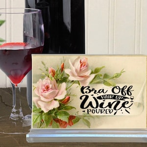 May include: A white sign with a floral background and the text "Bra Off Hair Up Wine Poured".