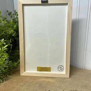May include: A framed print with a white background and a wooden frame. The print is approximately 8 inches by 11 3/4 inches. The frame has a small metal hanger on the back. The print has a gold label with the text "Nature's Glow" and a black and white circle with the letters "N.G."