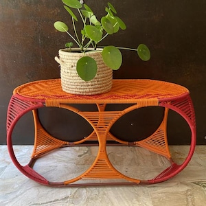 Peut inclure: Une table d'appoint en osier orange et rouge avec un plateau rond et deux ouvertures circulaires dans le piétement. Une plante en pot avec des feuilles vertes est posée sur la table.