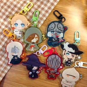 May include: A selection of acrylic keyrings with anime-style character designs. Each keyring showcases a unique character with different outfits and accessories, attached to a metal clasp. The keyrings are in various colours, including yellow, green, and red.
