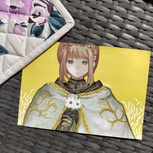 May include: A yellow art print featuring an anime-style illustration of a person with light brown hair and a white and gold cloak. The artwork includes a white flower and gold accents. A floral patterned pot holder is in the upper left corner.