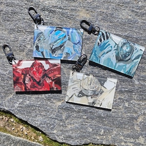 Könnte beinhalten: Vier rechteckige Schlüsselanhänger mit schwarzen Verschlüssen. Jeder Schlüsselanhänger zeigt eine andere abstrakte Illustration eines Roboters oder einer futuristischen Figur. Die Schlüsselanhänger sind in Rot-, Blau- und Grautönen gehalten, einer mit dem Text "HEY BUDDY".