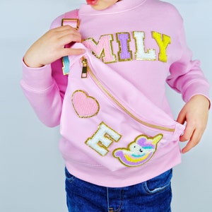 May include: A pink fanny pack with a gold zipper and a heart, letter "M" and unicorn patch. The fanny pack is worn by a child wearing a pink sweatshirt with the word "MILY" on the front and blue jeans.