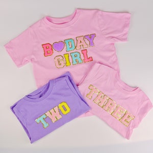 May include: Three pink and purple t-shirts with gold glitter lettering. The first shirt says "B'DAY GIRL" in rainbow colours. The second shirt says "TWO" in yellow and blue. The third shirt says "THREE" in gold.