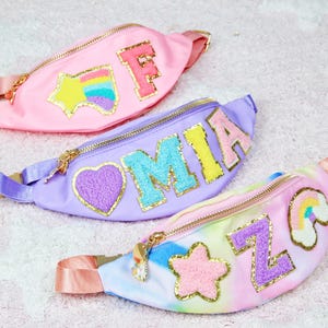 May include: Three colourful fanny packs with different designs. The first is pink with a rainbow and star, the second is purple with a heart and the letters MIA, and the third is tie-dye with a rainbow, star, and the letter Z.