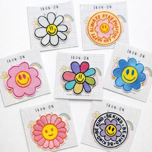 Può includere: Collezione di toppe termoadesive con disegni di fiori sorridenti in vari colori. Una toppa recita "Always Stay Positive", e un'altra dice "Be Kind". Ogni toppa è su un supporto a motivo a griglia.